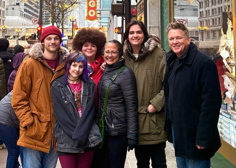 A group of six smiling people of various ages and styles poses together on a bustling Chicago sidewalk during the day.