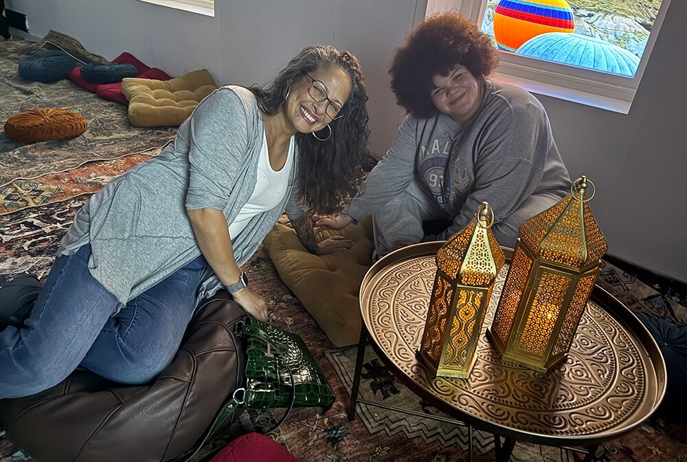 Two women smile while sitting on a patterned rug in a cozy, bohemian-style room.