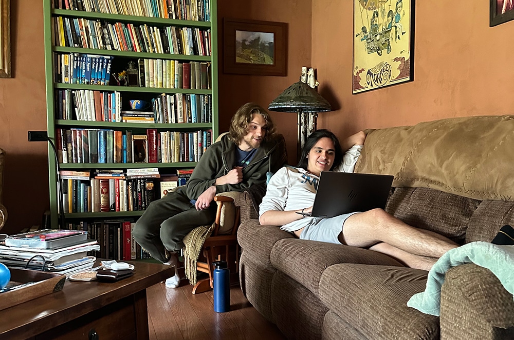 Two young men relax in a warm, earthy-toned living room with terracotta walls and hardwood floors.